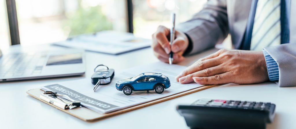 Close-up of a person signing a car financing agreement at a desk with a model car, keys, and calculator, representing vehicle purchase paperwork.