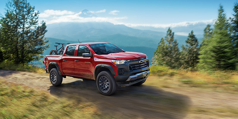 Red Chevrolet Colorado pickup truck driving on a forest road with mountains in the background