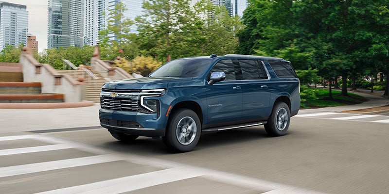 Blue Chevrolet Suburban SUV driving through a city street with trees and buildings in the background.